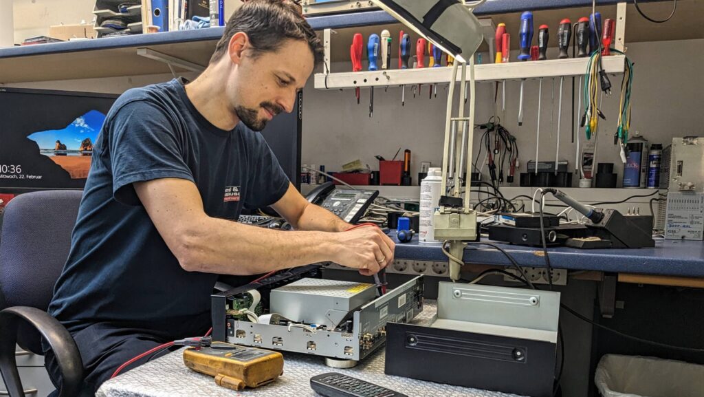 Ein Techniker prüft und repariert ein geöffnetes Elektronikgerät an einer Werkbank mit Messgerät und Werkzeug.