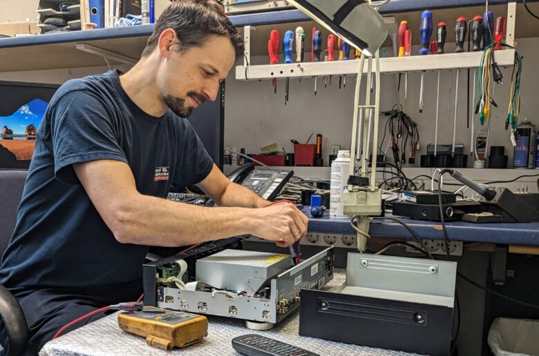 Ein Techniker prüft und repariert ein geöffnetes Elektronikgerät an einer Werkbank mit Messgerät und Werkzeug.
