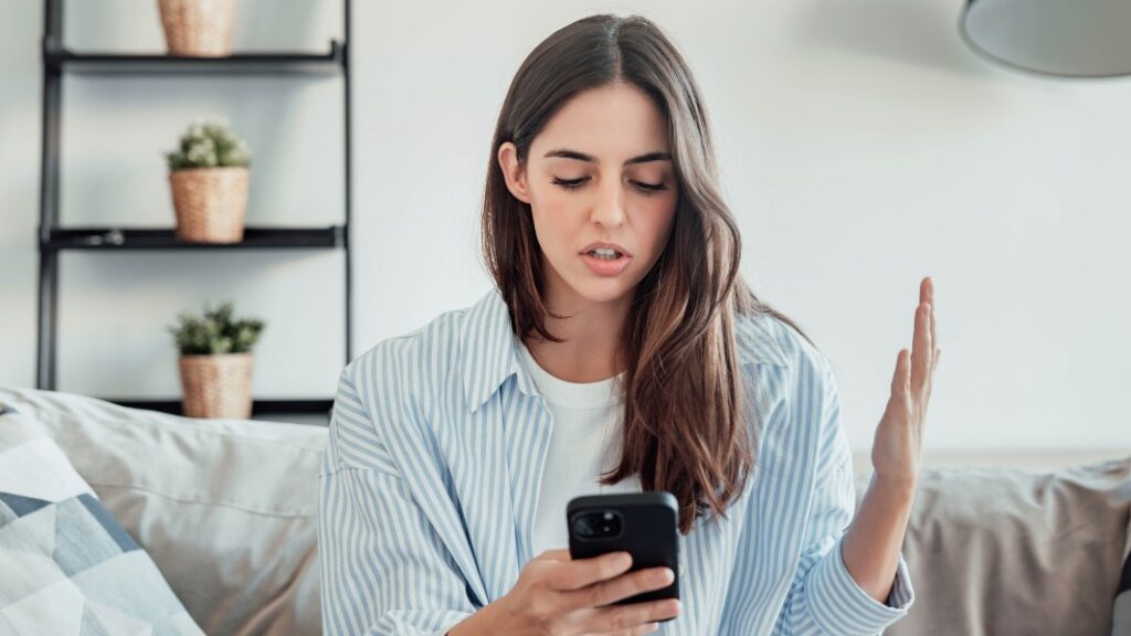 Gerätekompatibilität: Eine Frau sitzt auf einem Sofa und blickt irritiert auf ihr Smartphone, während sie eine Hand ratlos hebt, als Symbol für Probleme mit Smart-Home-Apps.