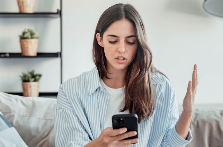 Gerätekompatibilität: Eine Frau sitzt auf einem Sofa und blickt irritiert auf ihr Smartphone, während sie eine Hand ratlos hebt, als Symbol für Probleme mit Smart-Home-Apps.