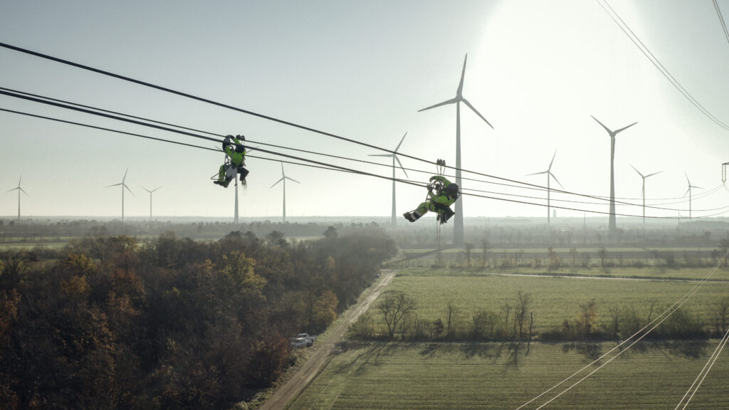 Stromkosten: Zwei Monteure arbeiten an einer Hochspannungsleitung über einer Landschaft mit Windkraftanlagen.