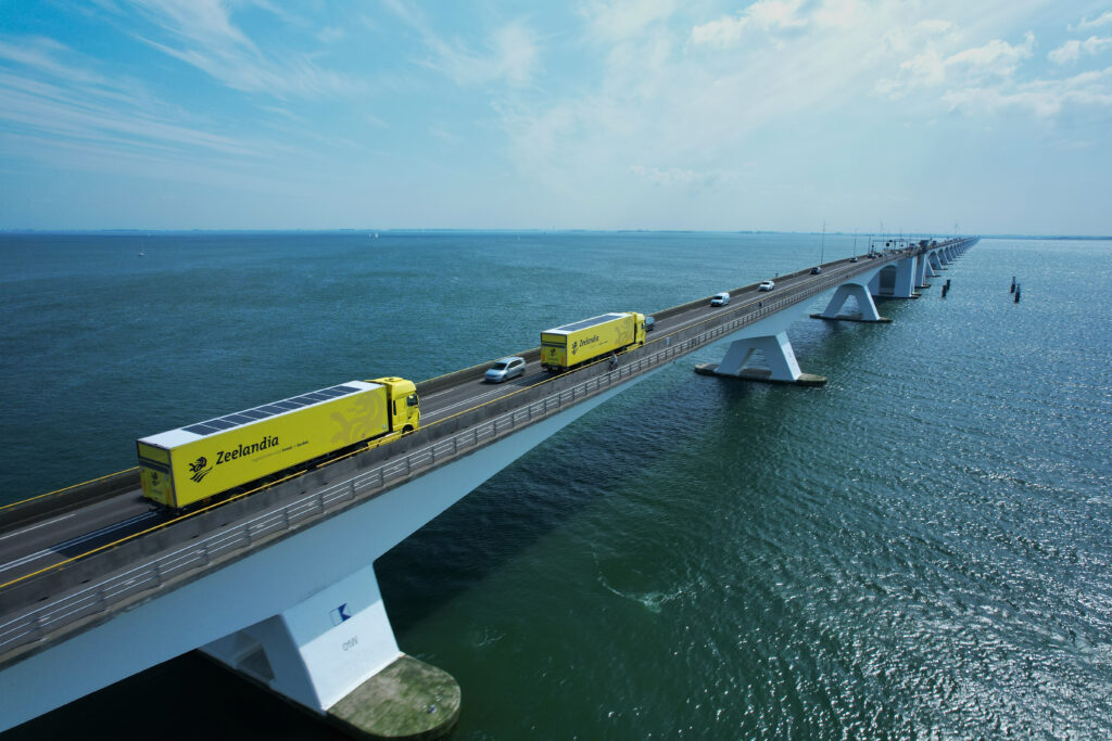 Zwei gelbe Lkw mit Solarmodulen auf den Aufliegern fahren über eine lange Brücke über das Meer.