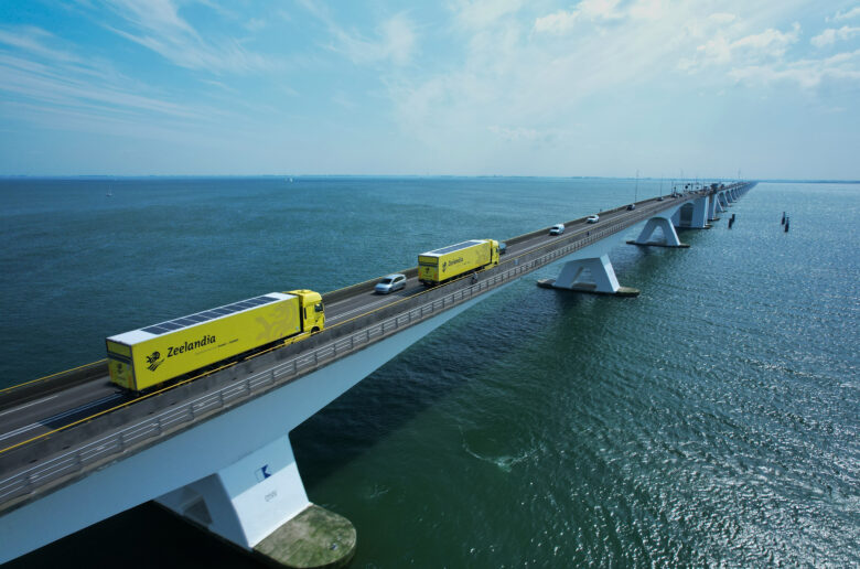 Zwei gelbe Lkw mit Solarmodulen auf den Aufliegern fahren über eine lange Brücke über das Meer.