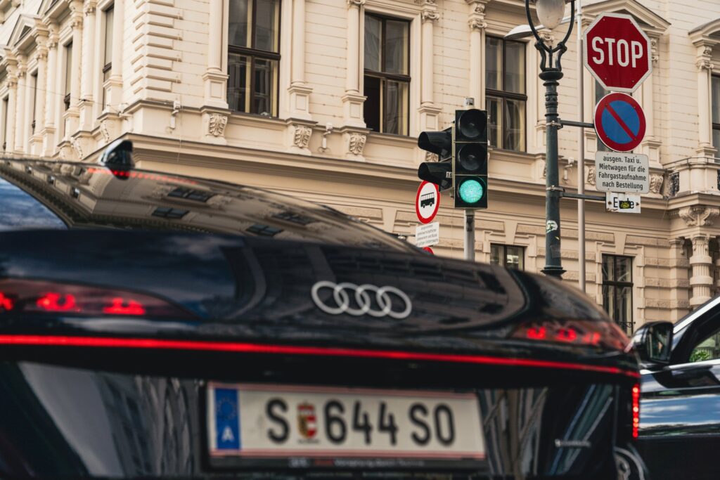 Heck einer dunklen Limousine mit österreichischem Kennzeichen vor einer grün leuchtenden Verkehrsampel, einem Stoppschild und einer historischen Hausfassade.