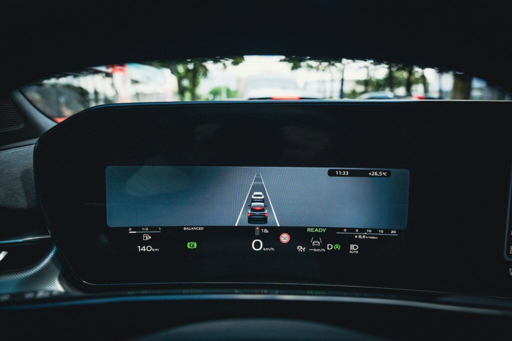 Fahrerdisplay eines stehenden Autos mit 0 km/h und einem roten Ampelsymbol samt Countdown von 18 Sekunden; durch die Windschutzscheibe ist der Stadtverkehr zu sehen.