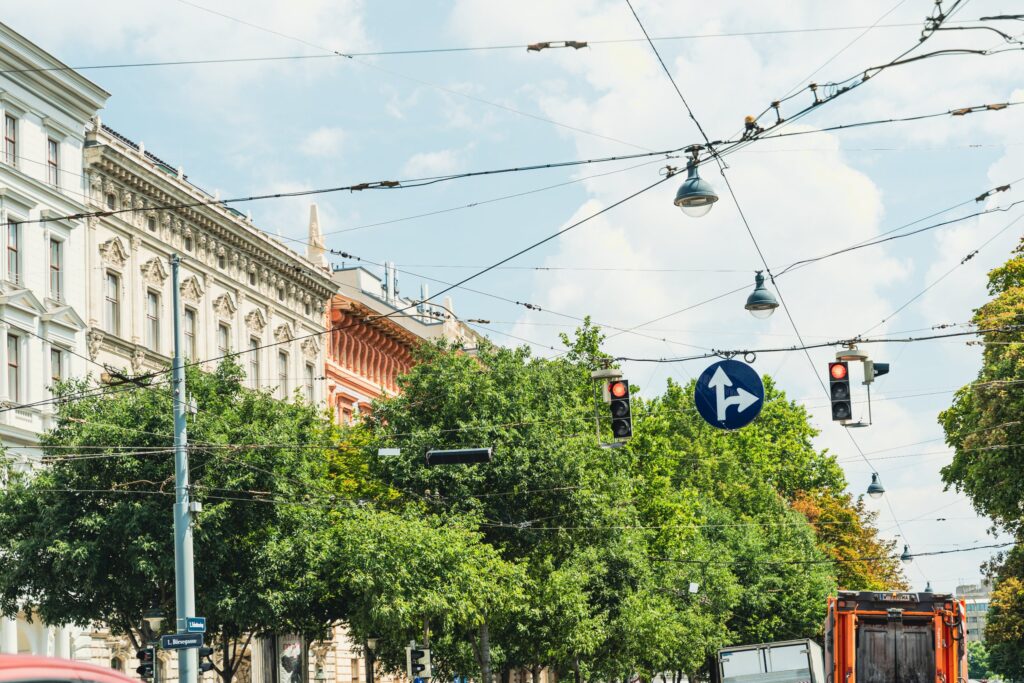 Zwei rot leuchtende Verkehrsampeln und ein blaues Gebotszeichen hängen über einer baumgesäumten Straße in der Wiener Innenstadt; links ein Straßenschild „Börsegasse“.
